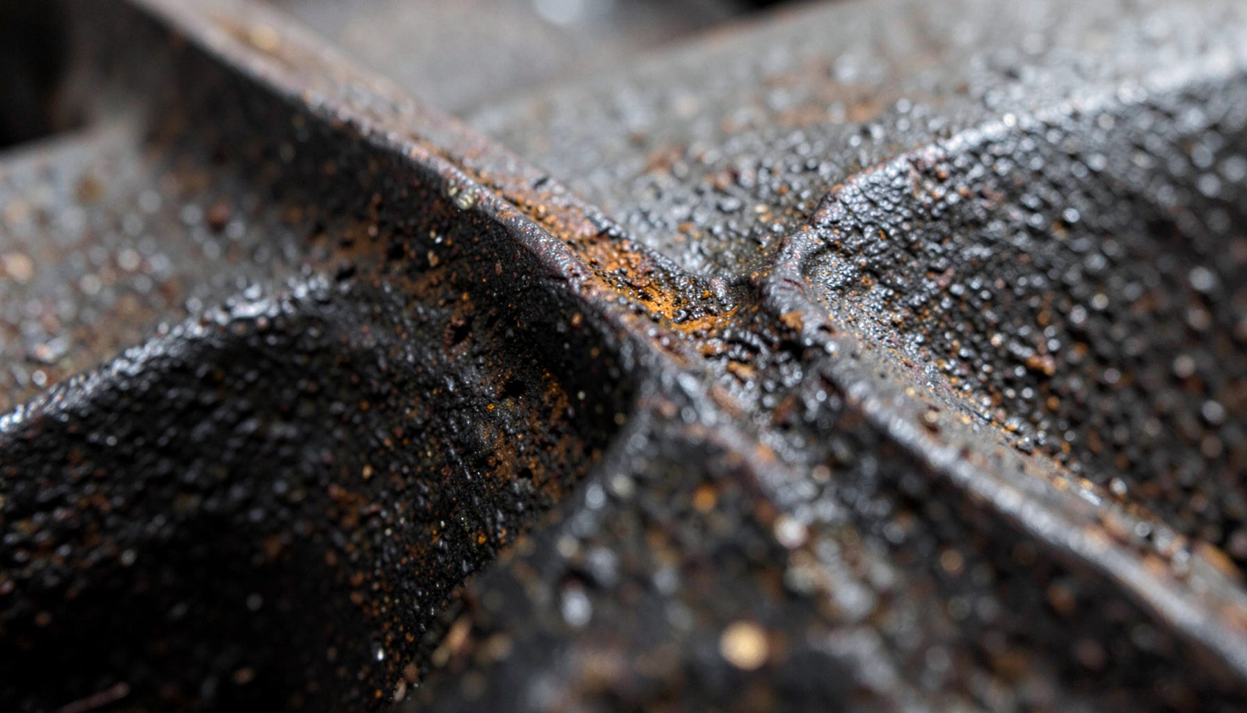 Extreme close-up of aged cast iron, its dark grey surface marked by fine pitting and a faint bloom of amber rust around a machined edge, conveying decades of quiet endurance.